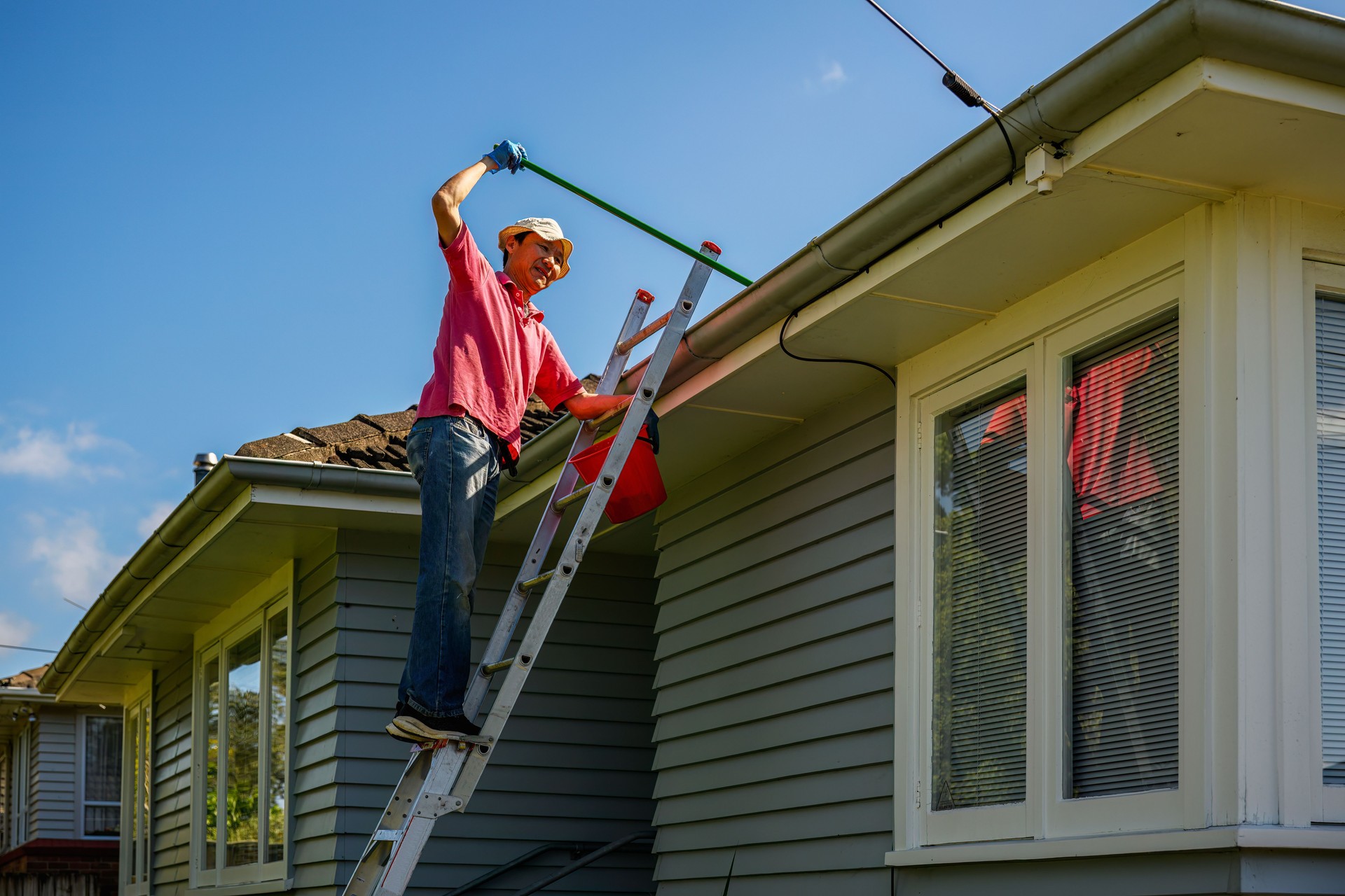 Man standing on the ladder and cleaning gutter with a gutter cleaning shovel. Home maintenance work. Auckland.